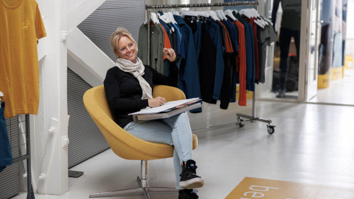 Christine Haraldstad-Sindum sitzt in einem Sessel mit Block und Stift. Neben ihr ist eine Kleiderstange aufgestellt.