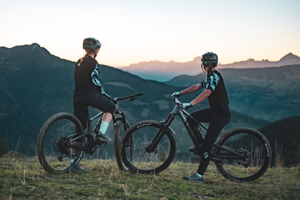 Zwei Fahrradfahrerinnen sitzen auf Mointainbikes und blicken in die Ferne. Dort sind Berge im Hintergrund zu sehen.