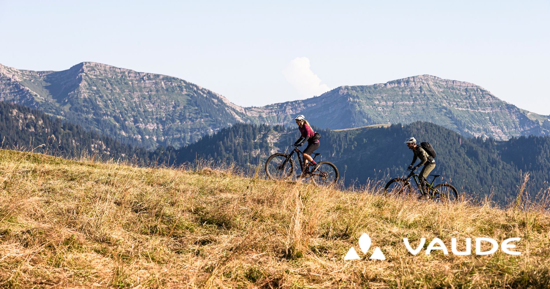 2 Personen fahren mit Mountainbikes über eine Wiese einen Berg hoch.