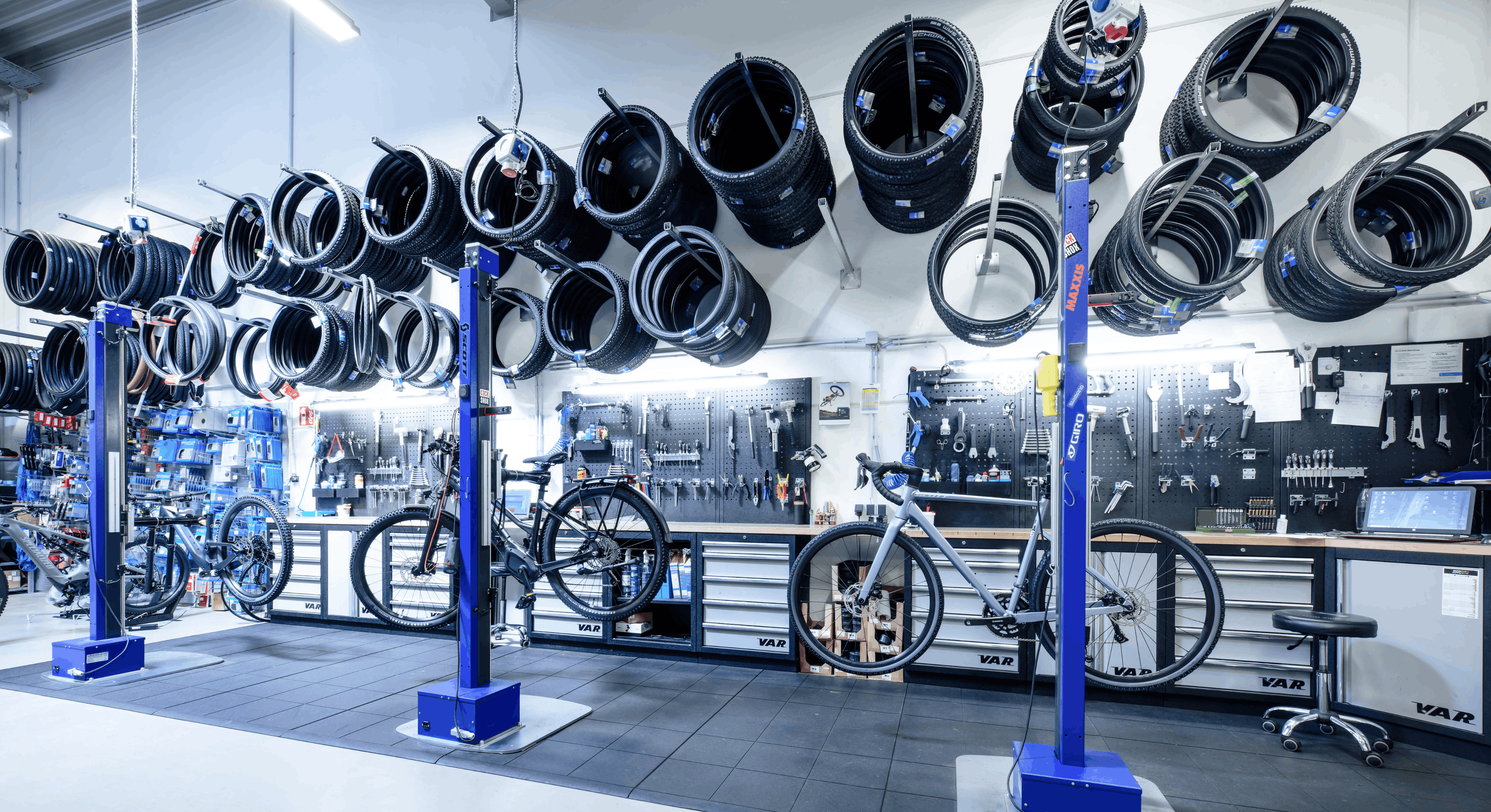Fahrradwerkstatt mit drei Arbeitsplätzen an denen jeweils ein Fahrrad hängt. Große Arbeitsfläche mit Werkzeuen und von obern hängen neue Fahrradreifen